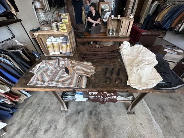 Retail table at Brass Tacks Provisions with folded shirts, accessories, grooming products, and clothing racks
