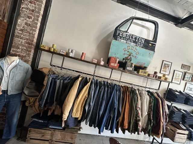 Clothing rack with jackets and shirts at Brass Tacks Provisions beneath a vintage Pendleton Round-Up sign