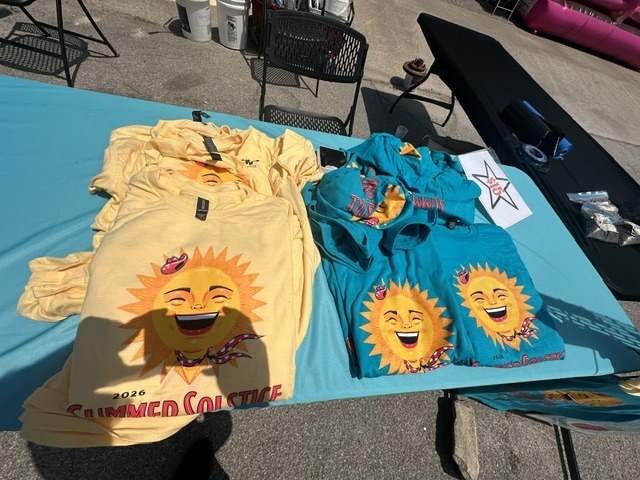 Colorful 2026 Summer Solstice shirts with smiling sun designs displayed on a table.