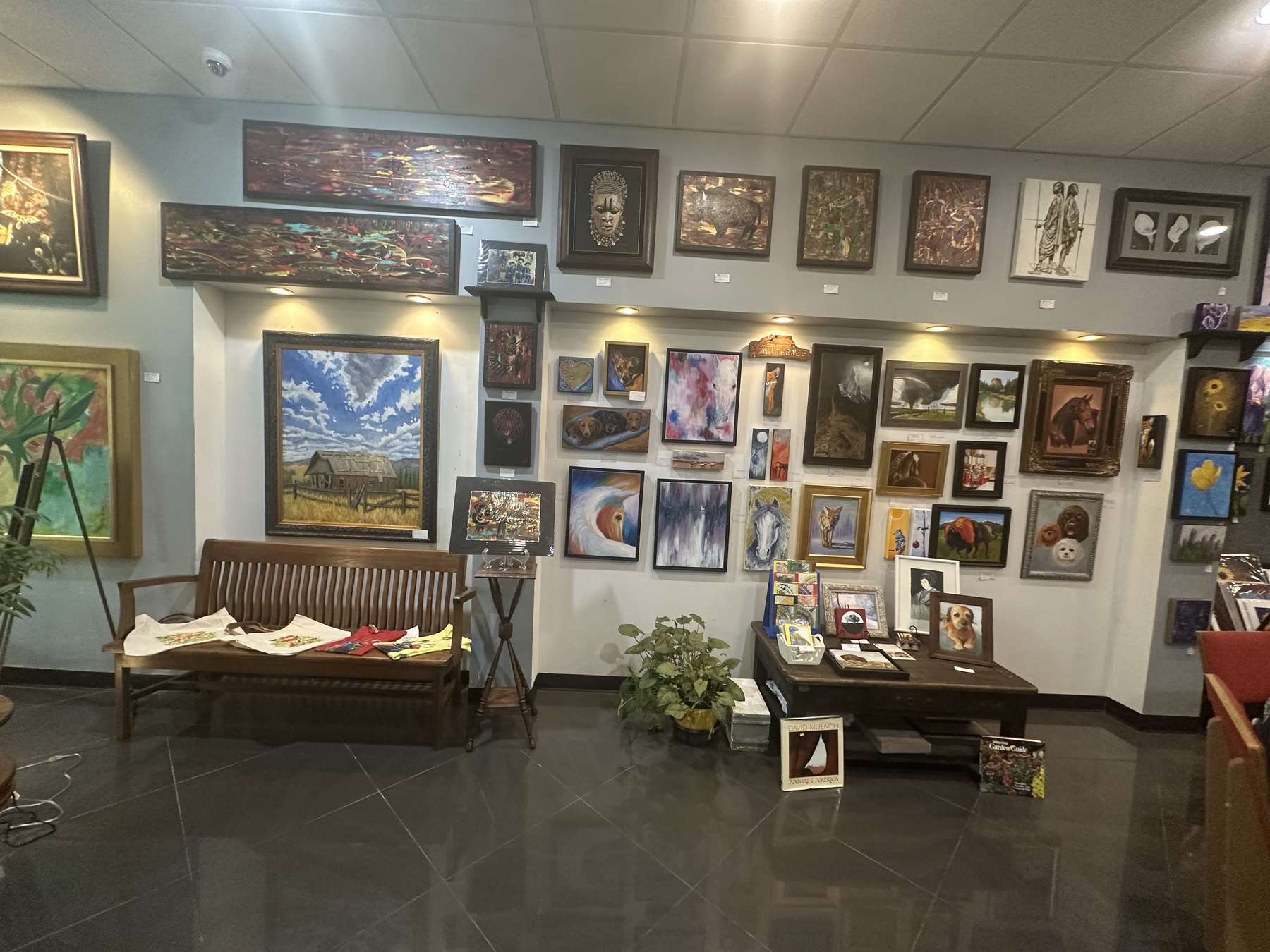 Interior wall of Art Central Gallery filled with framed paintings, small artworks, a bench, and display tables