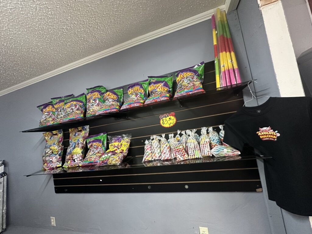 Candy bags, wrapped treats, colorful candy sticks, and an Axolotl Dulceria T-shirt on a wall display.
