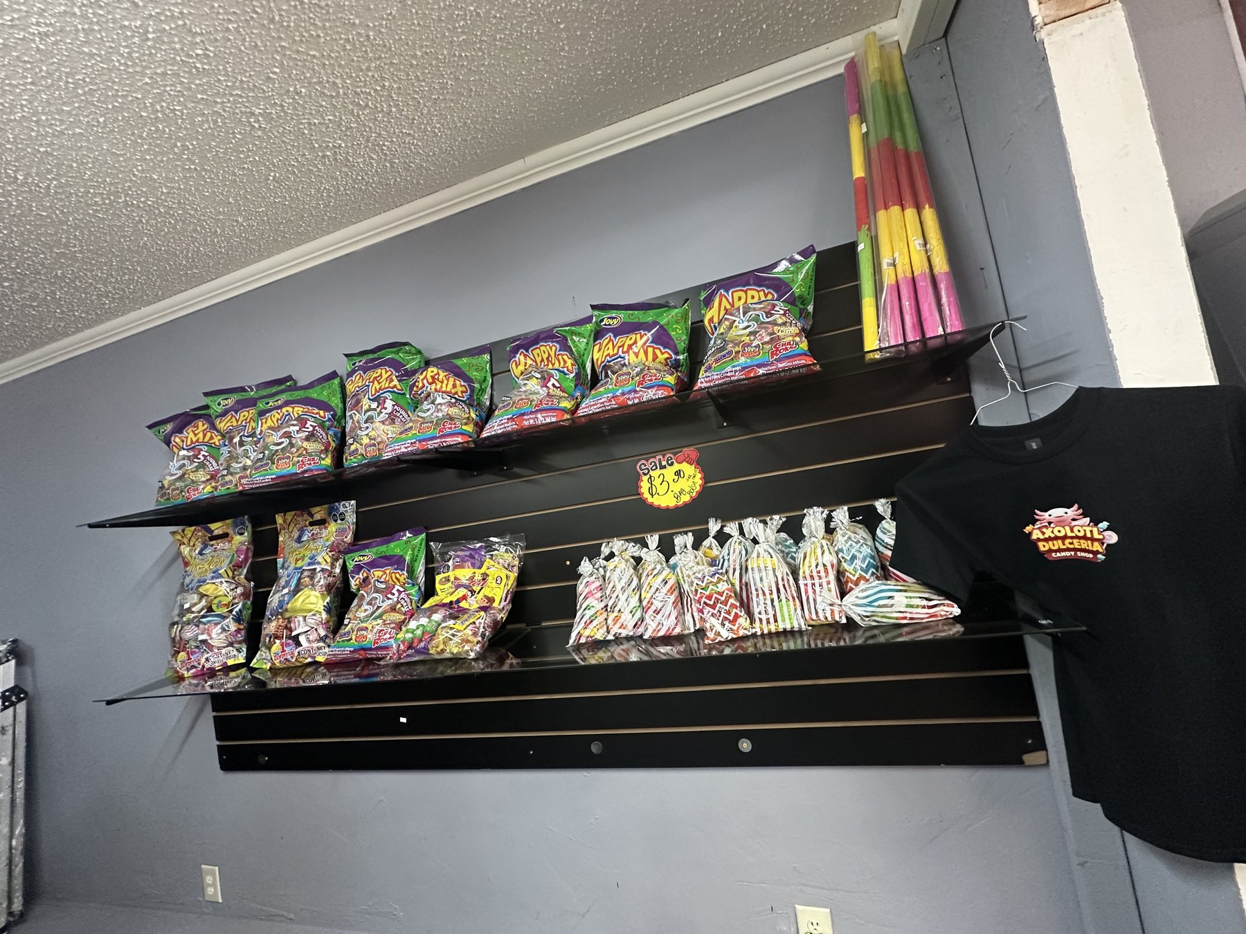 Candy bags, wrapped treats, colorful candy sticks, and an Axolotl Dulceria T-shirt on a wall display.