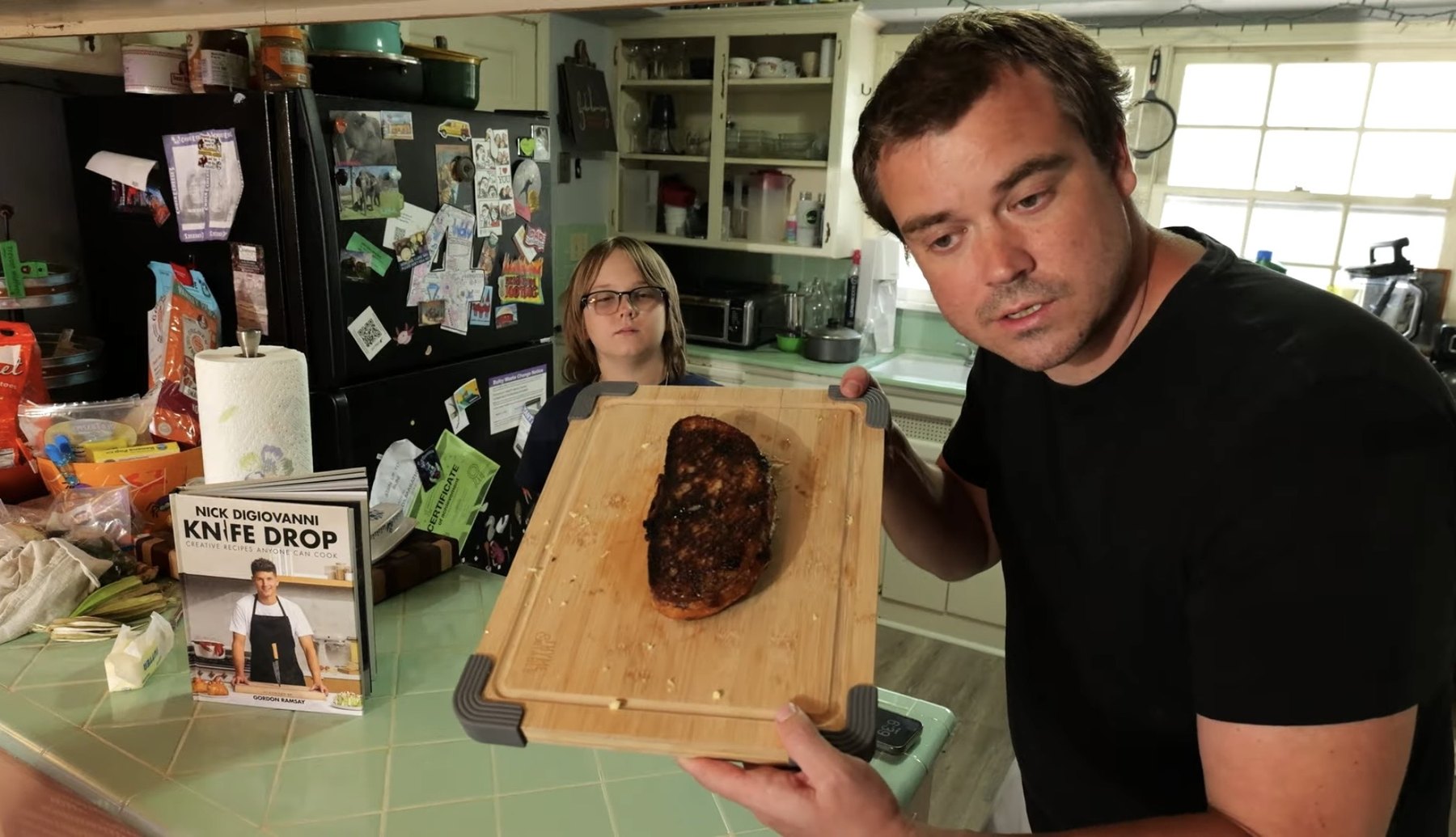 Adult holds a cutting board with a toasted blueberry brie grilled cheese beside the Knife Drop cookbook.
