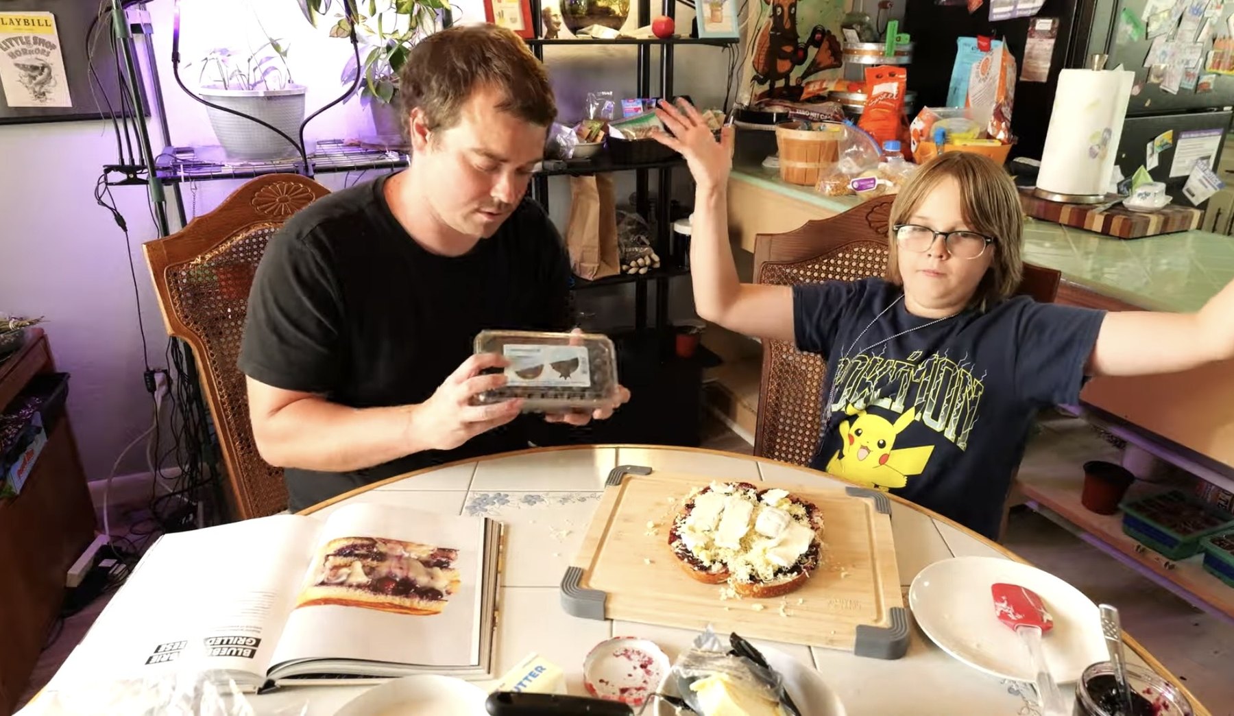 Adult and child assemble blueberry brie grilled cheese at a table with the Knife Drop cookbook open nearby.