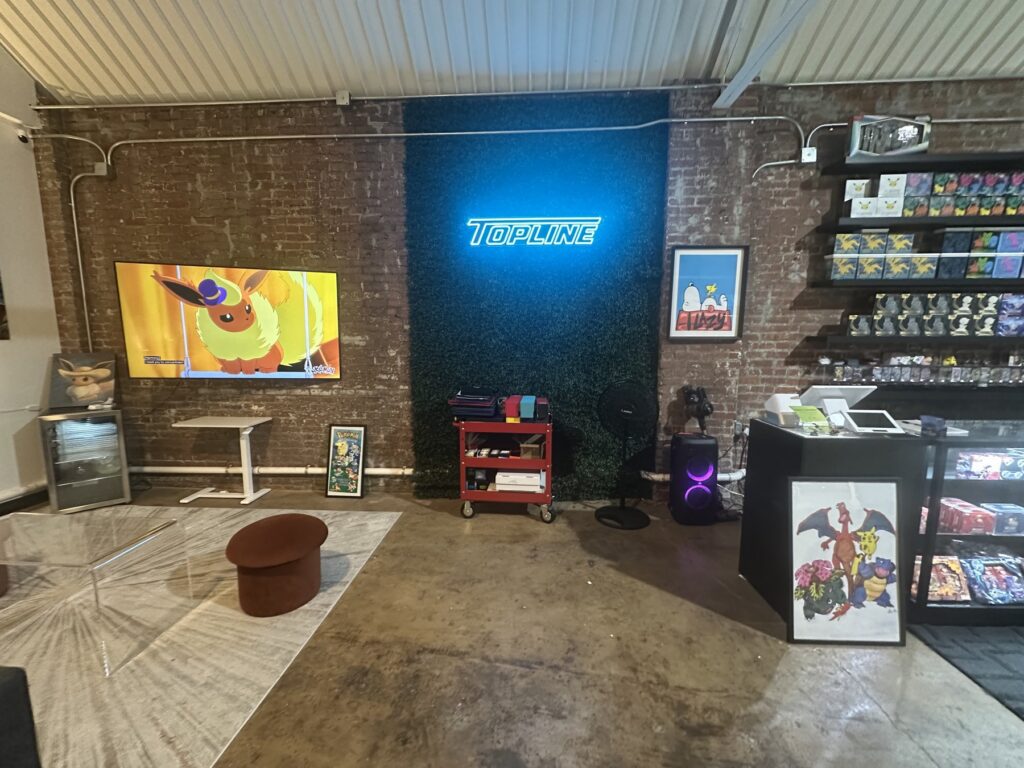 Interior of Topline Collectibles with a blue Topline neon sign, exposed brick wall, Pokemon artwork, and display cases.