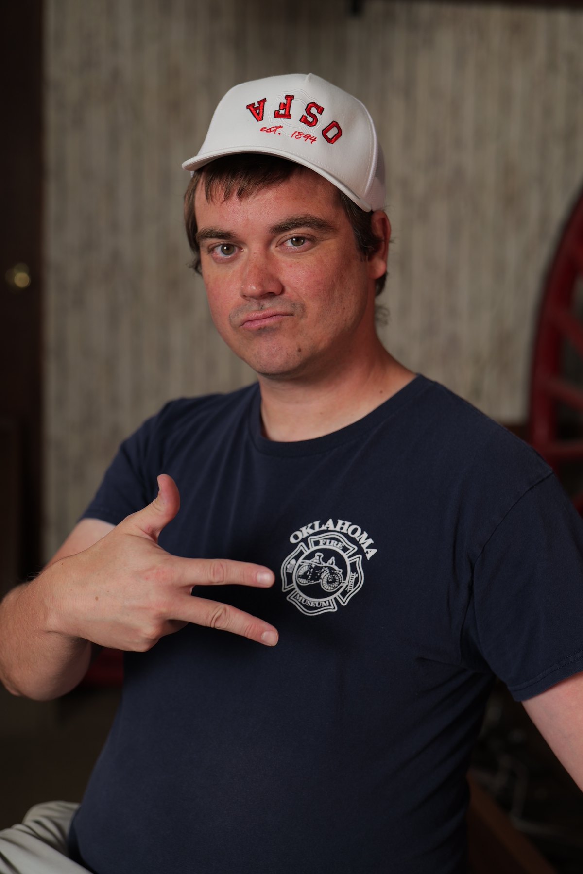 Participant wearing an Oklahoma Fire Museum shirt and white OSFA cap points toward the museum logo.
