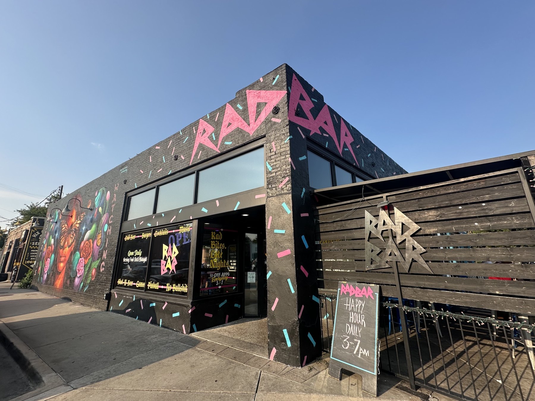 Exterior of Rad Bar in Oklahoma with pink RADBAR lettering, colorful murals, patio fencing, and a happy hour sign.
