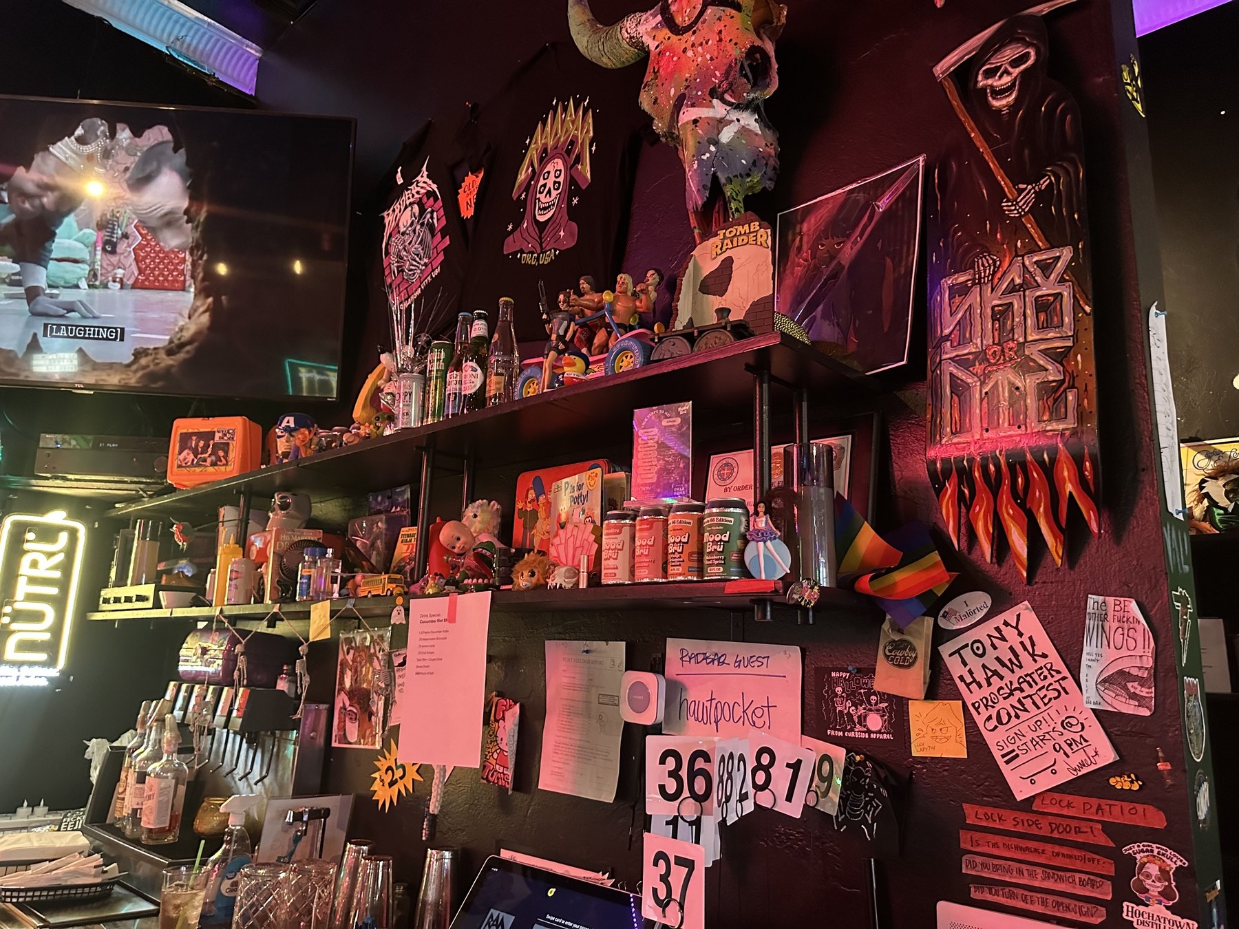Decorated wall behind the bar at Rad Bar with shelves, toys, flyers, skull art, stickers, and colorful signs.