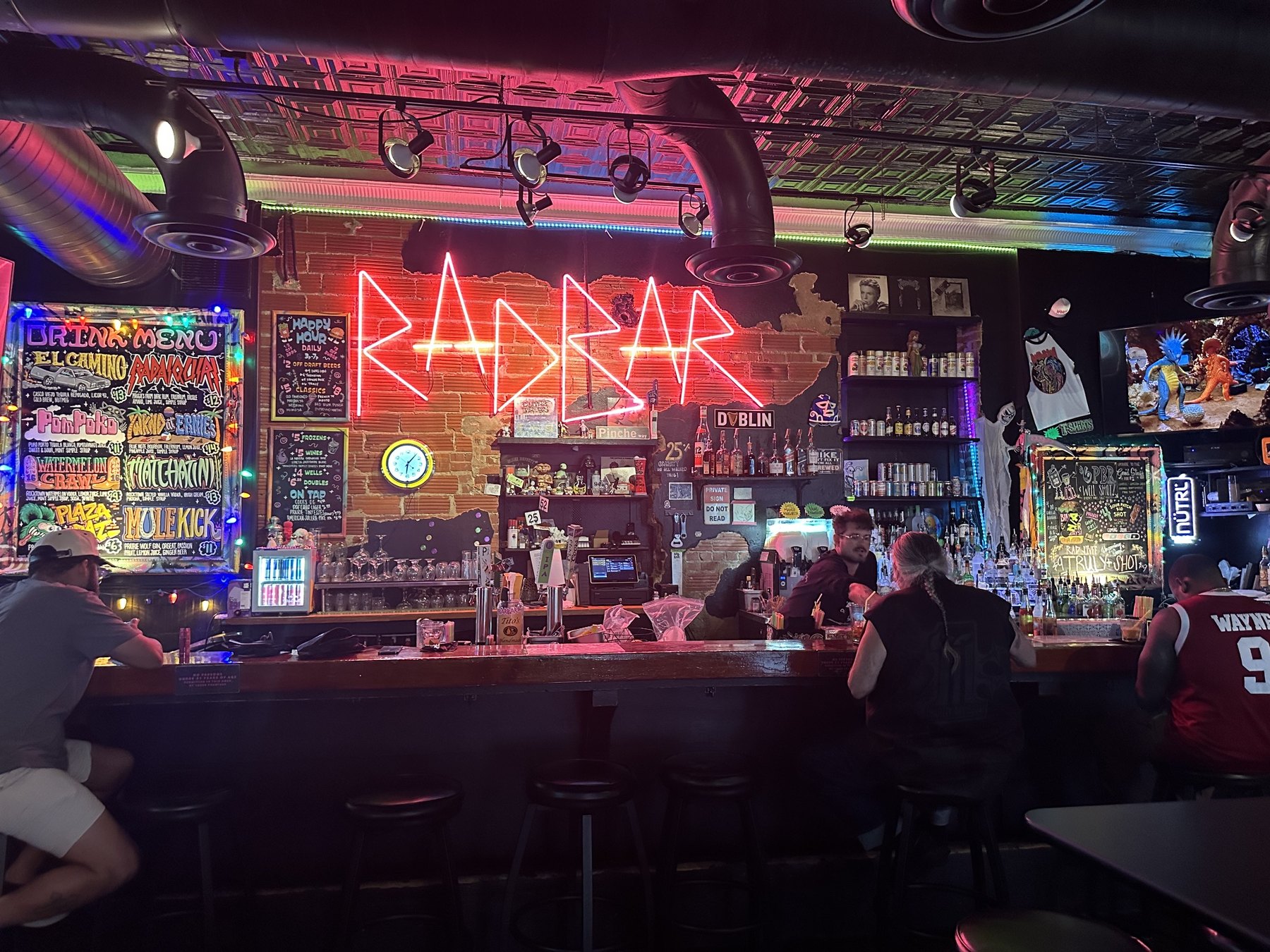 Interior of Rad Bar with a large red neon RADBAR sign above the bar, colorful menu boards, patrons, and moody lighting.