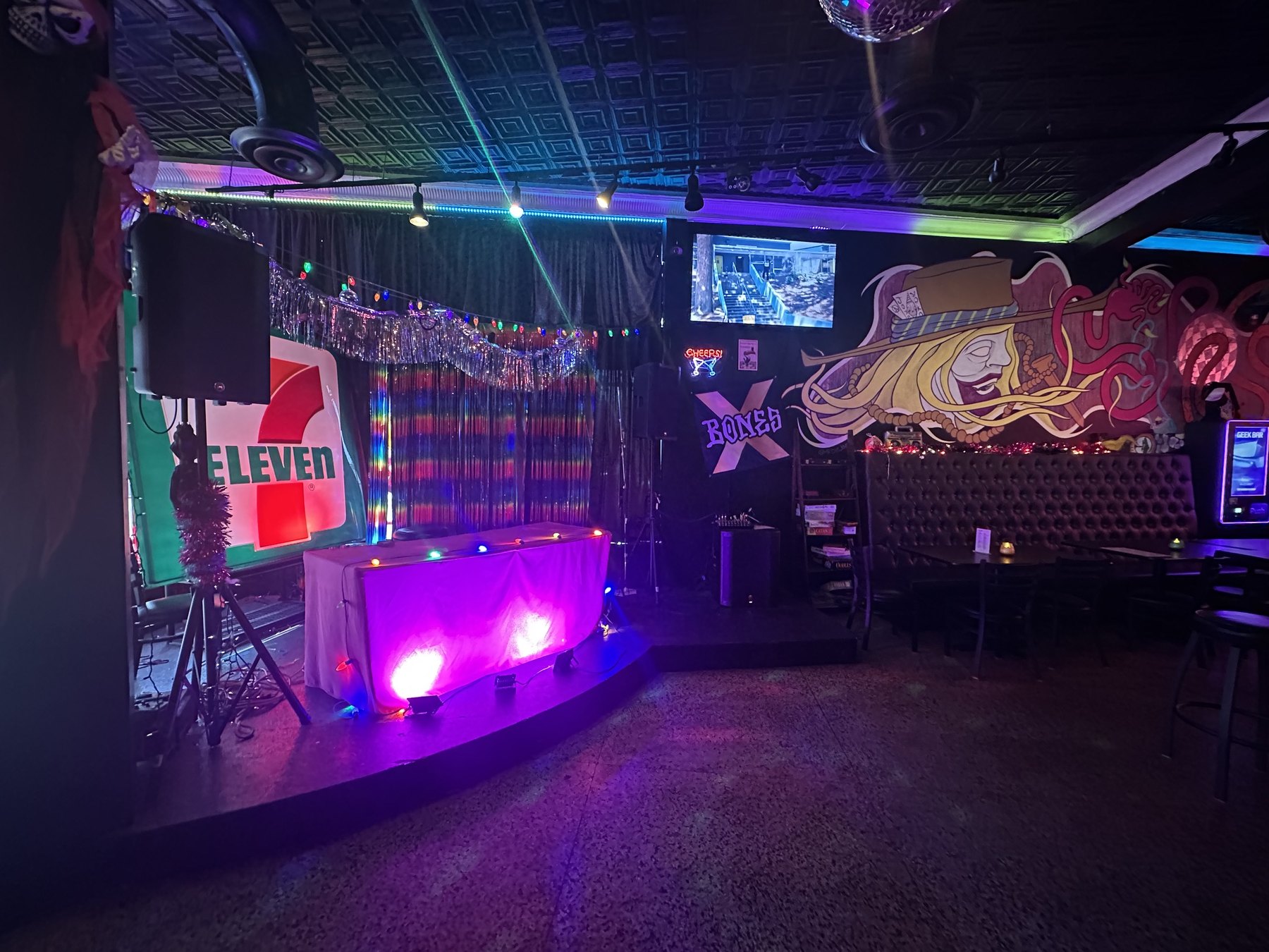 Small stage and DJ area at Rad Bar with colorful lights, a 7-Eleven sign, murals, booths, and a disco ball.