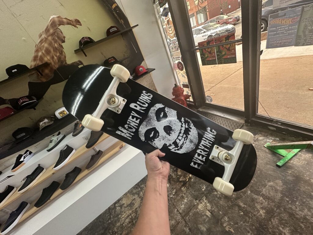 A completed black Money Ruins Everything skateboard with white wheels being held inside an Oklahoma City skate shop.