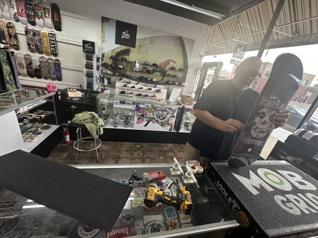 A shop worker at Money Ruins Everything holds a skateboard deck at the counter with tools and parts nearby.