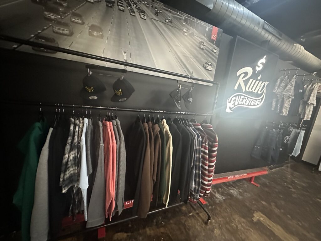 Clothing rack inside Money Ruins Everything below a large wall graphic and illuminated shop logo.