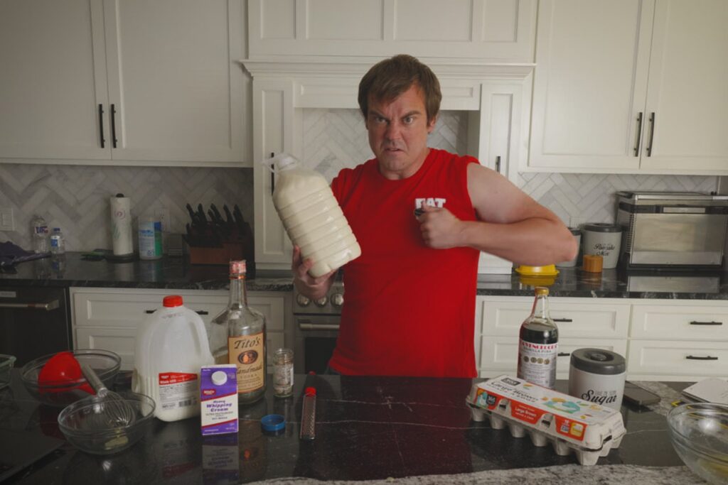 Cook holding a large jug of homemade Fight Milk beside milk, vodka, eggs, cream, sugar, and vanilla on a kitchen counter.