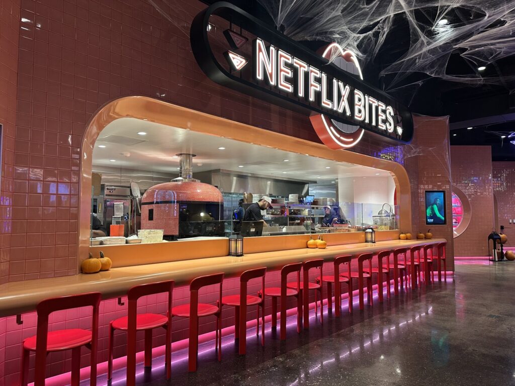 Netflix Bites Dallas counter with red stools, pink tile, neon sign, open kitchen, and Halloween web decorations