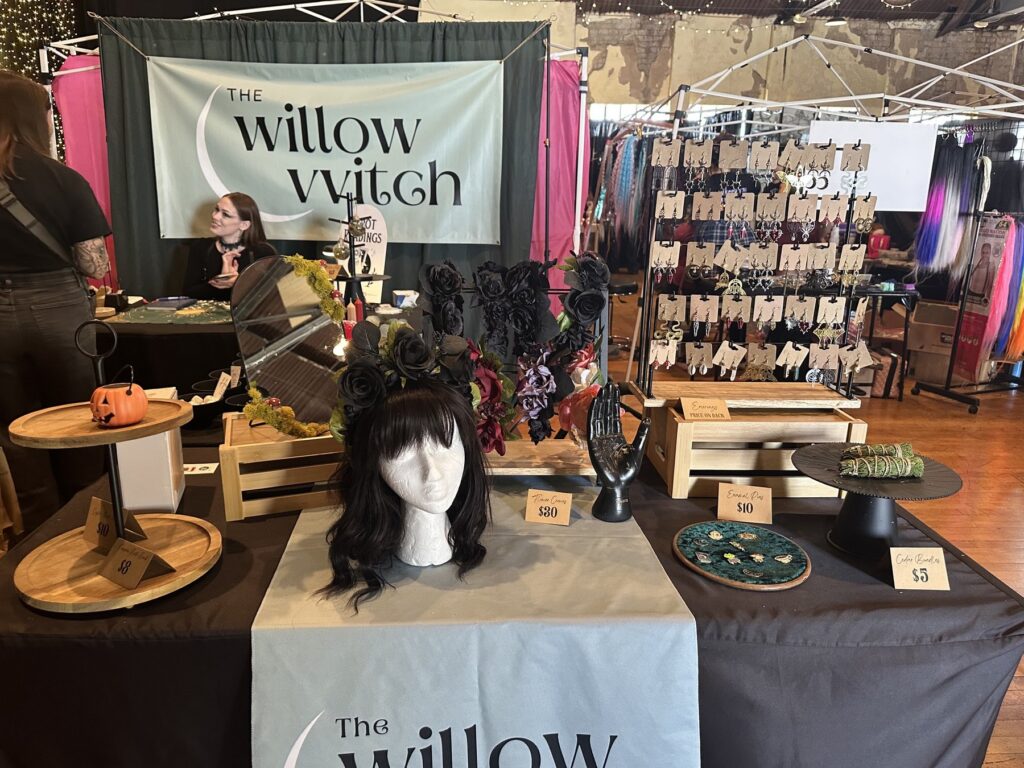 The Willow Witch booth with earrings, sage bundles, accessories, and black floral headwear at the Gothic Market Festival.