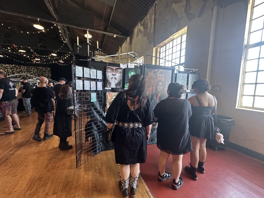 Festival attendees dressed in black looking at gothic art displays near tall windows.