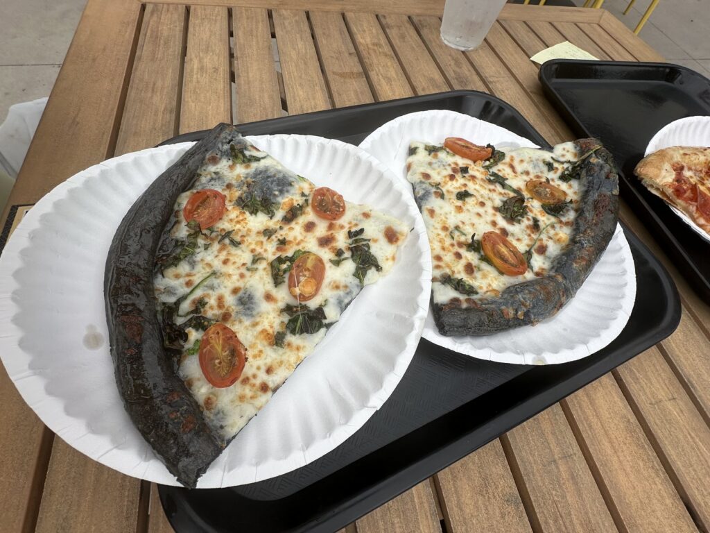 Two slices of black crust pizza with cheese, greens, and tomatoes from Eastside Pizza House