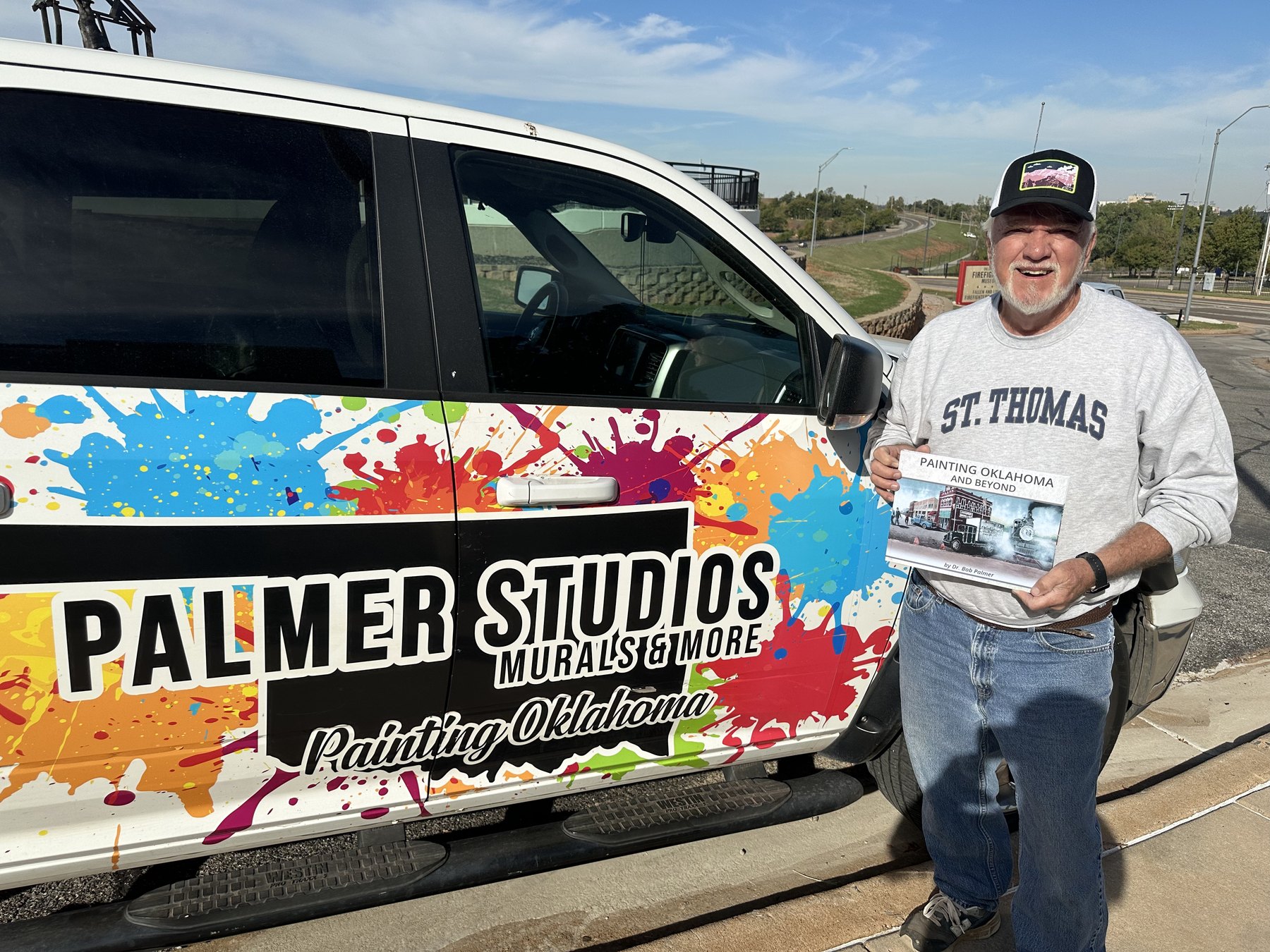 Dr. Bob Palmer holding Painting Oklahoma and Beyond beside the colorful Palmer Studios truck.