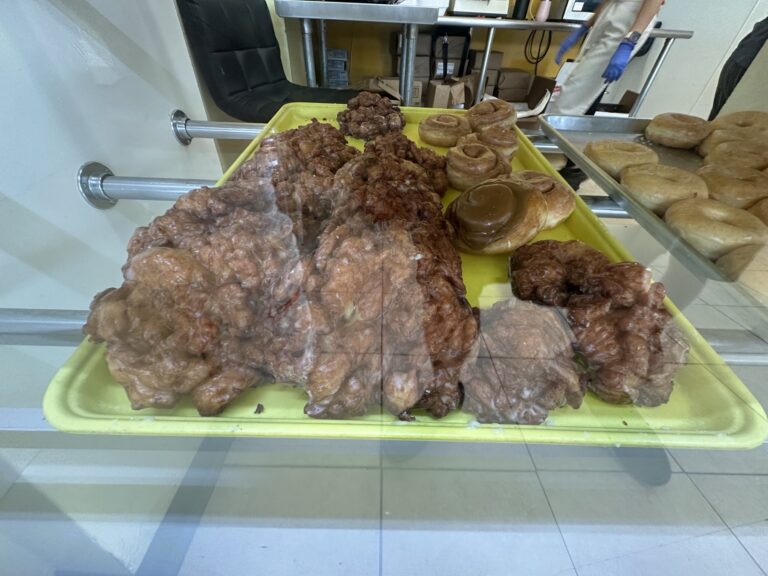 Large glazed apple fritters and cinnamon rolls displayed in a donut shop bakery case.