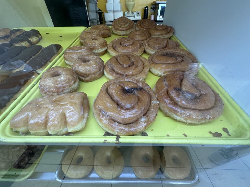 Glazed cinnamon rolls, glazed donuts, and a bear claw pastry in a donut shop display case.