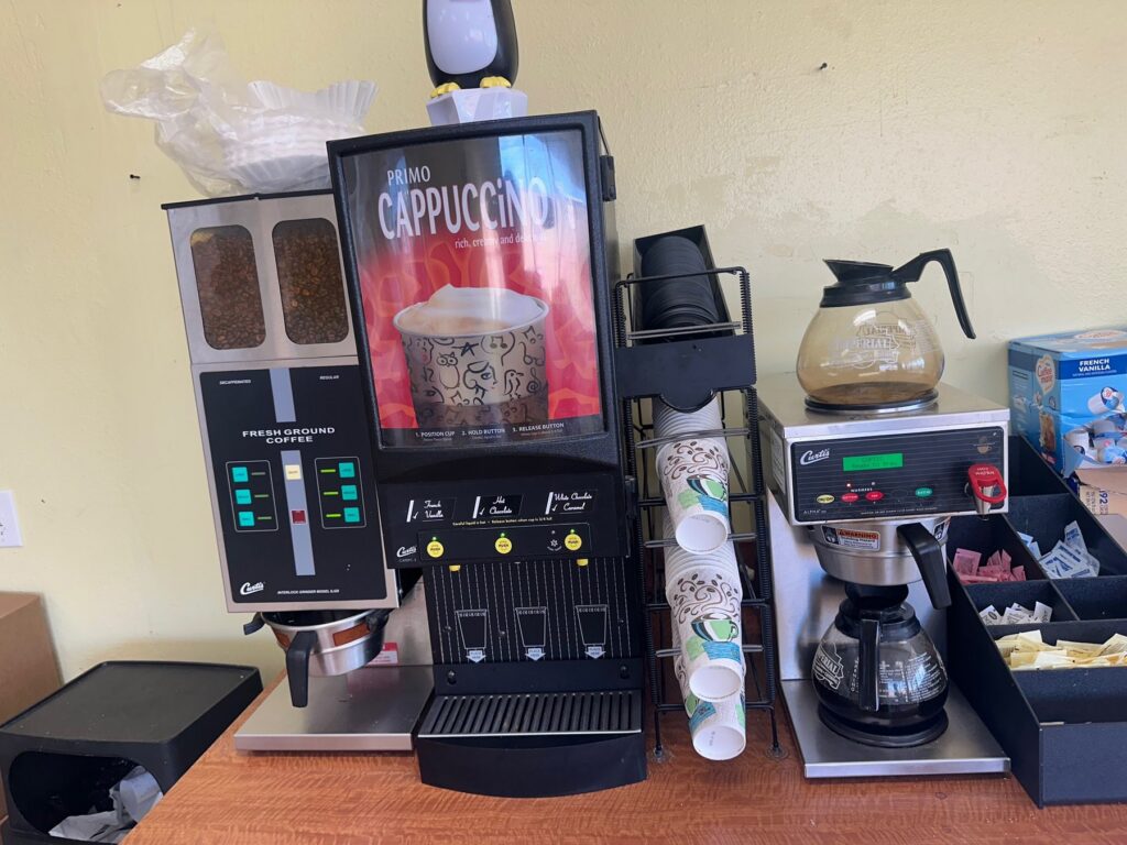 Self-serve coffee and cappuccino station with cups and condiments inside a donut shop.