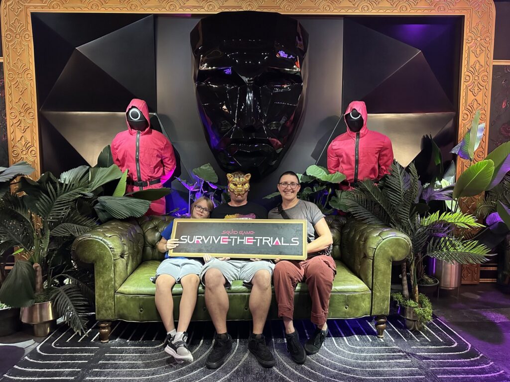Guests posing with a Squid Game: Survive the Trials sign at Netflix House Dallas, surrounded by pink guards and a large black mask display.