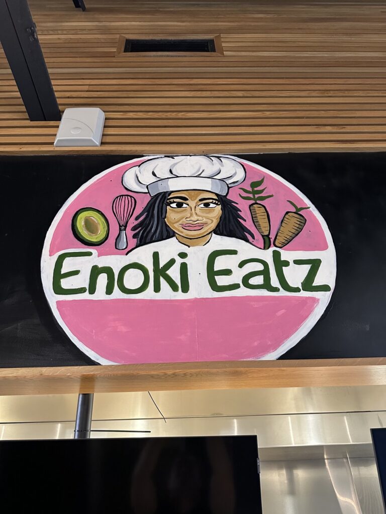 Close-up of the Enoki Eatz logo sign with a chef illustration and vegetables.
