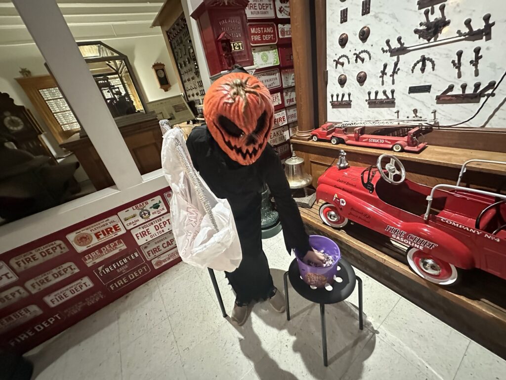Pumpkin-headed Halloween figure holding a white bag beside a fire-truck exhibit and candy bowl.