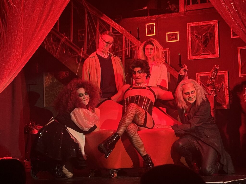 Cast members perform The Rocky Horror Picture Show on a red-lit stage with a gothic set and red chaise.