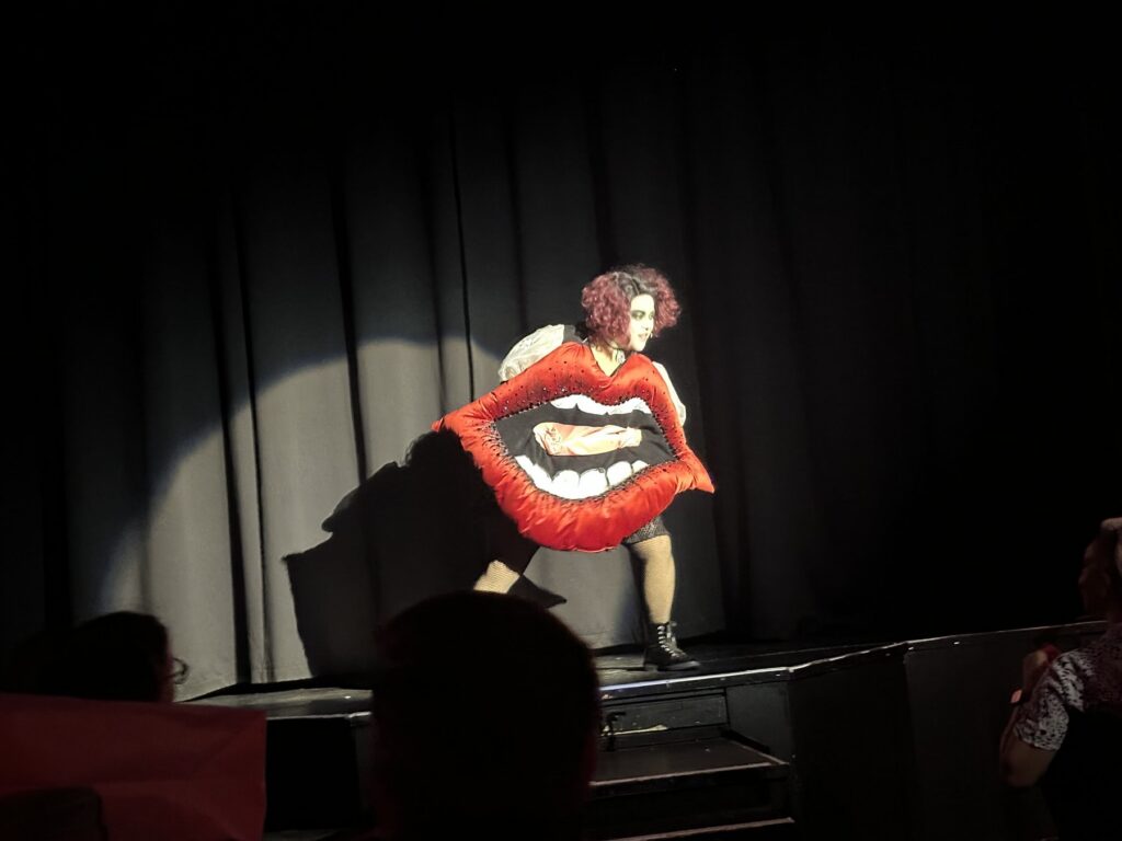 A Rocky Horror performer wears a large red lips costume while standing in a spotlight onstage.