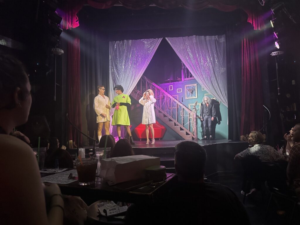 Audience members watch a Rocky Horror stage performance from tables in an intimate theater setting.