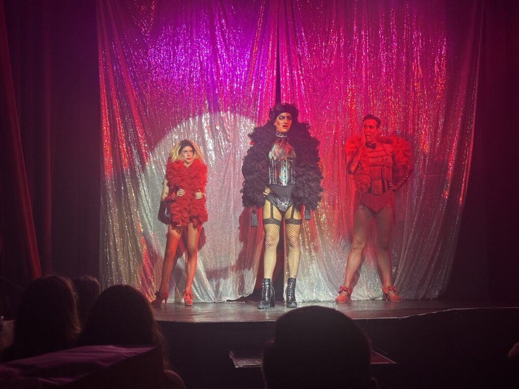 Three Rocky Horror performers pose in dramatic red and black costumes under stage lights.