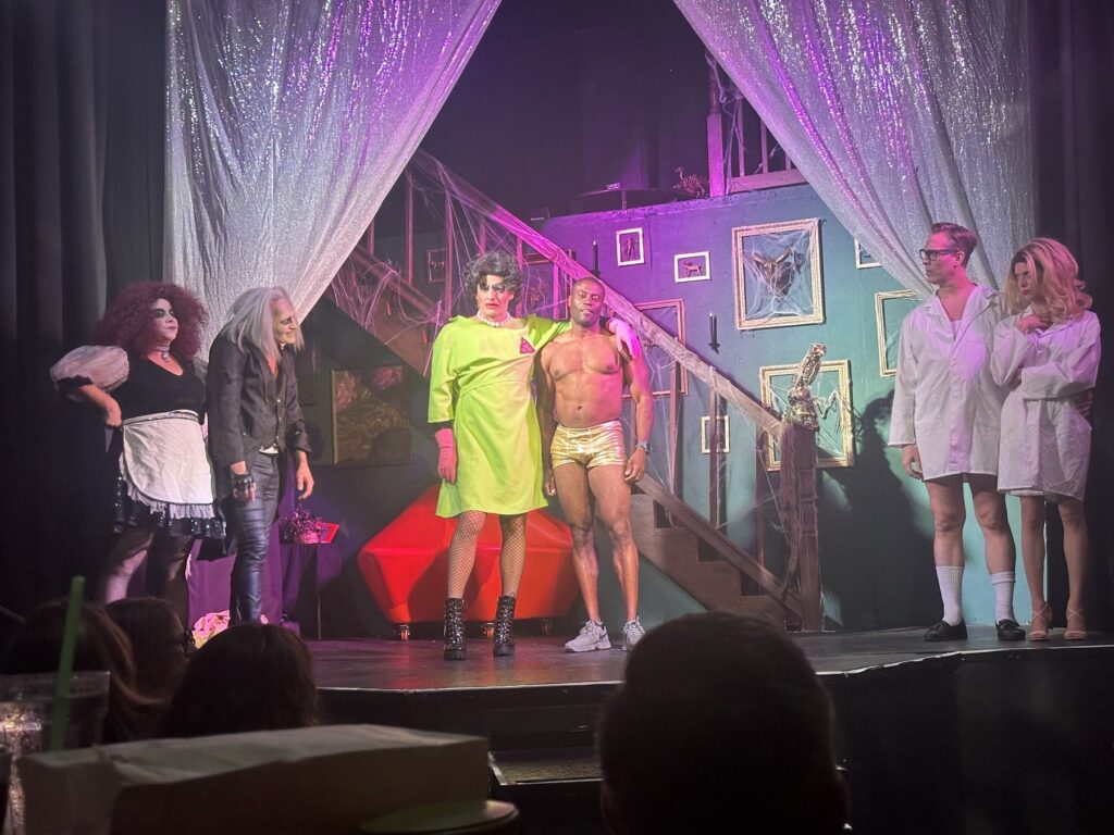 Several Rocky Horror cast members perform on a purple-lit stage with a staircase and cobwebbed set.