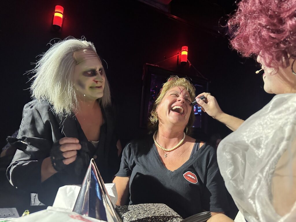 Costumed Rocky Horror performers interact with a laughing audience member and apply makeup during the show.