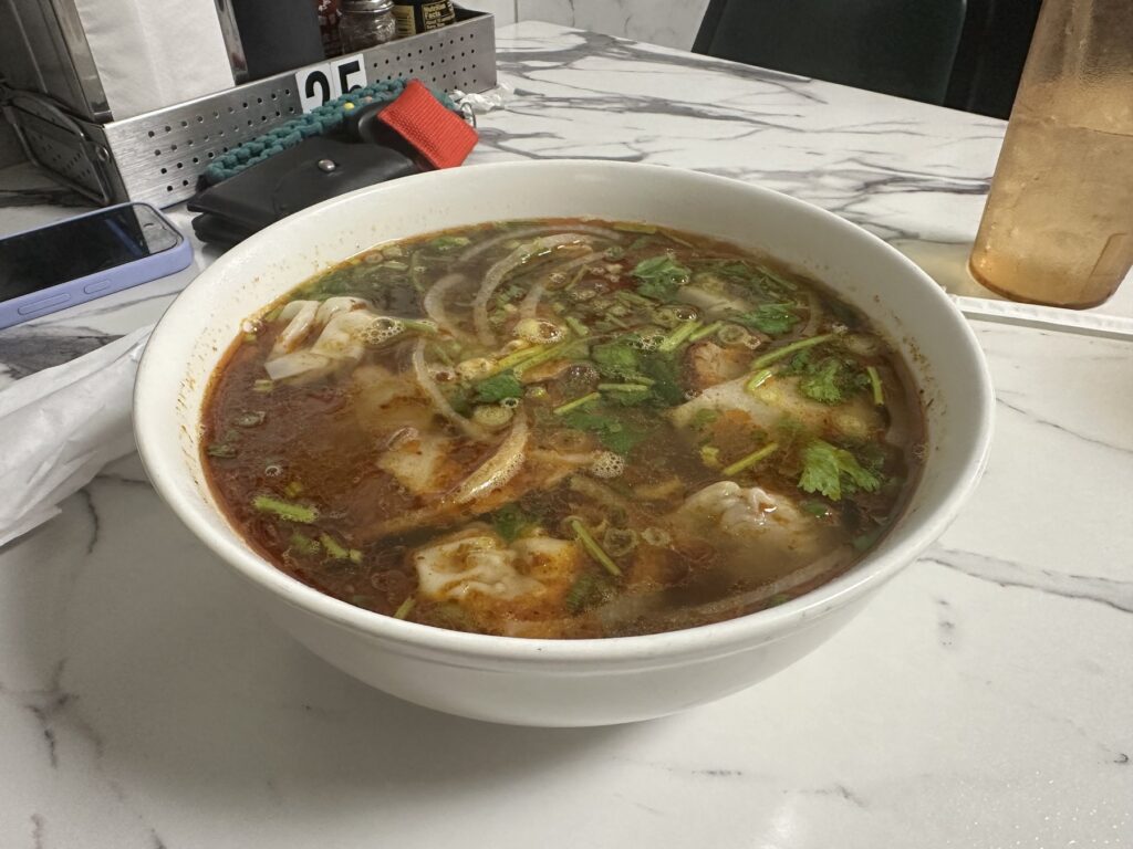 Close-up of a Vietnamese noodle soup bowl with reddish broth, herbs, onions, and meat at Pho Cuong.