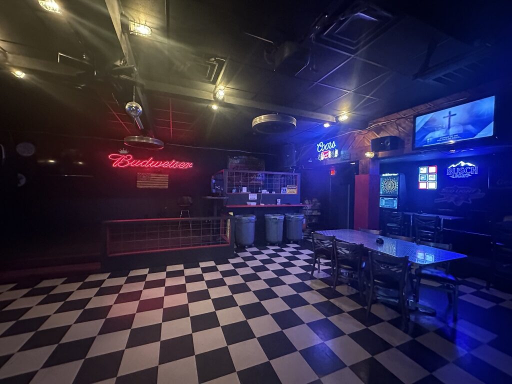 Dimly lit JR’s Pub and Grill interior with checkered floor, tables, TVs, and beer neon signs.